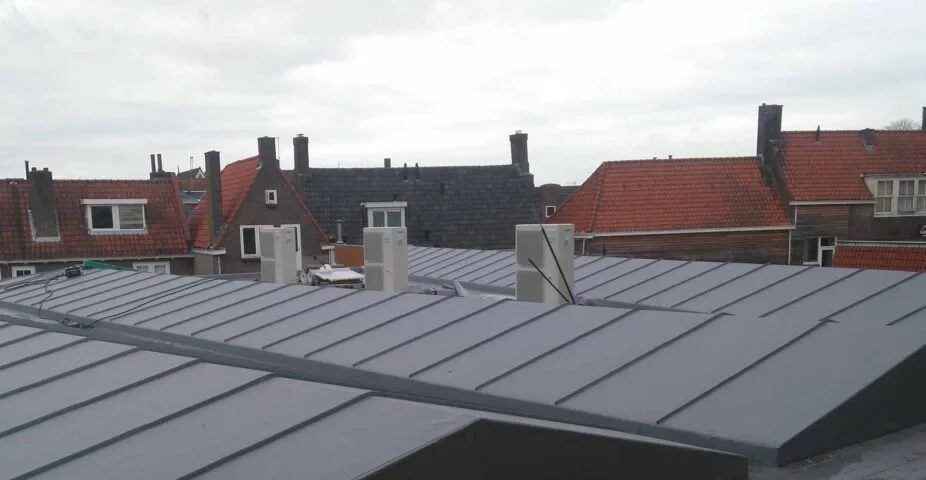 Grijs zinken dakvlak met staande naden, daarachter pannendaken